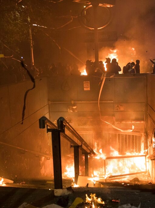 At night, you view stairs descending to a subway station on fire as people watch on around the perimeter wall.