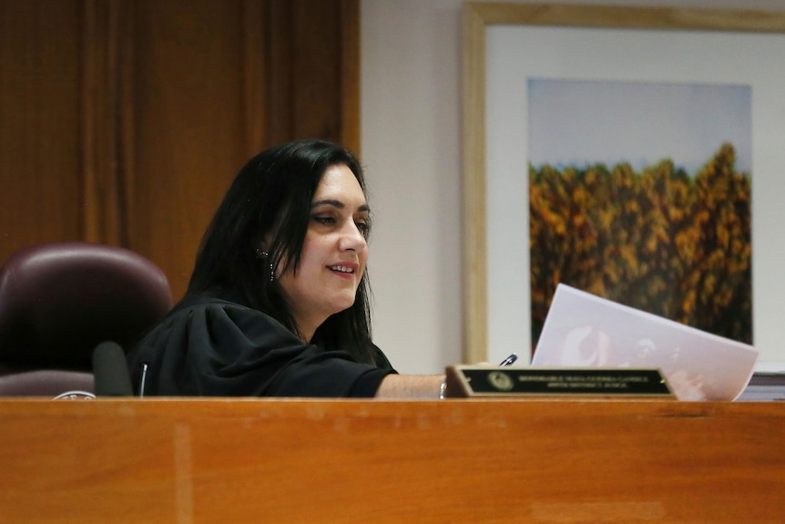 A female judge with dark hair sits in a courtroom examining a piece of paper and smiling slightly.