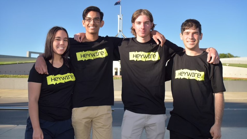 Four young people stand in front of parliament house in Heywire shirts with arms around each other