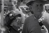 Black and white still from clip filmed in 1961, showing an elderly man and wife in hats on a busy street, talking to a reporter