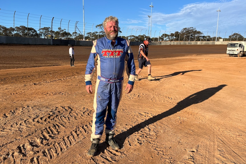 A man with white hair and a bear in racing coveralls on a dirt track