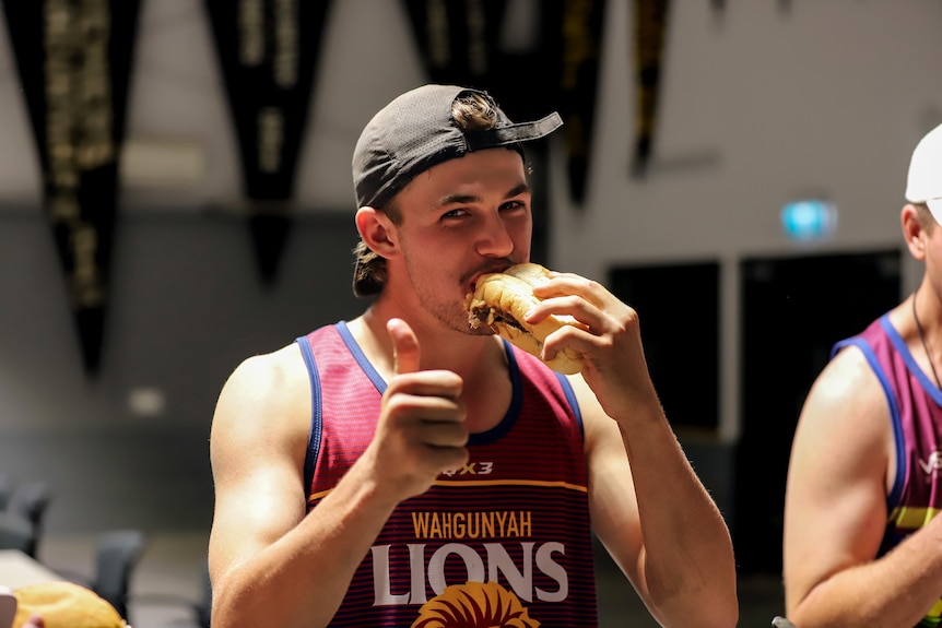 A young man wearing a singlet and shorts smiles while giving a thumbs up gesture while eating a lamb roll.