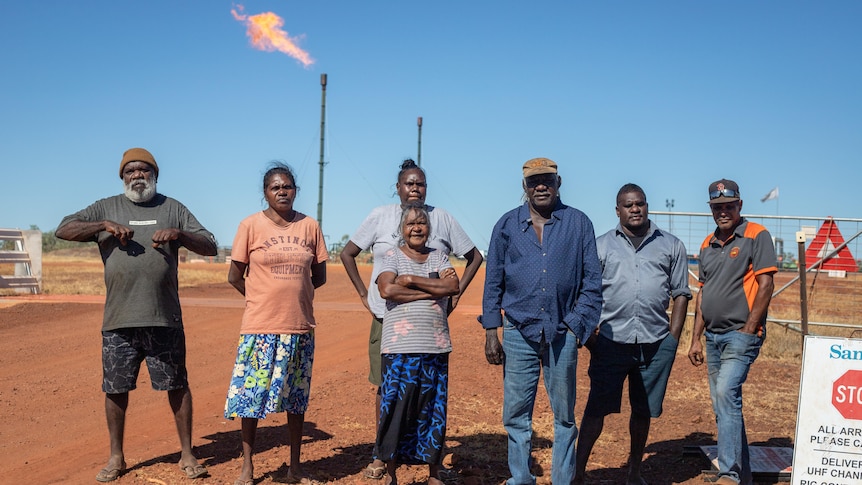 a group of Indigenous people standing in front of a flaring gas well.