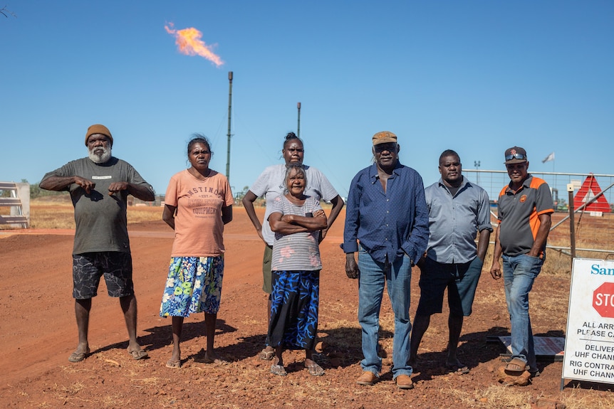 a group of Indigenous people standing in front of a flaring gas well.