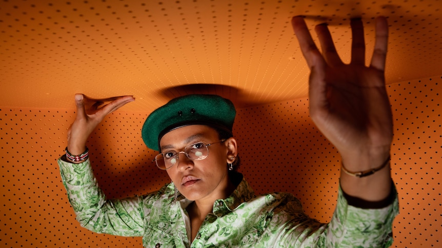 A woman stands in small bright orange room. Her fingers brush the ceiling and she wears a green beret and a serious expression.