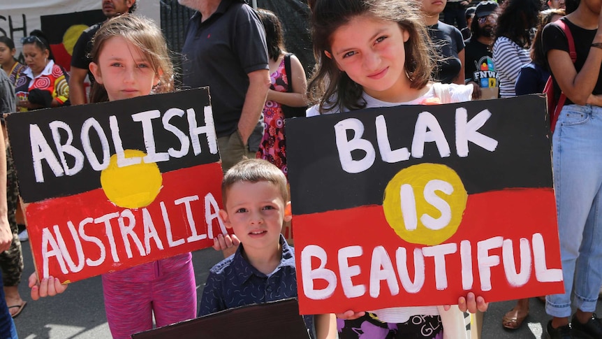 Three children, two holding indigenous flag signs which read "abolish Australia" and "black is beautiful".