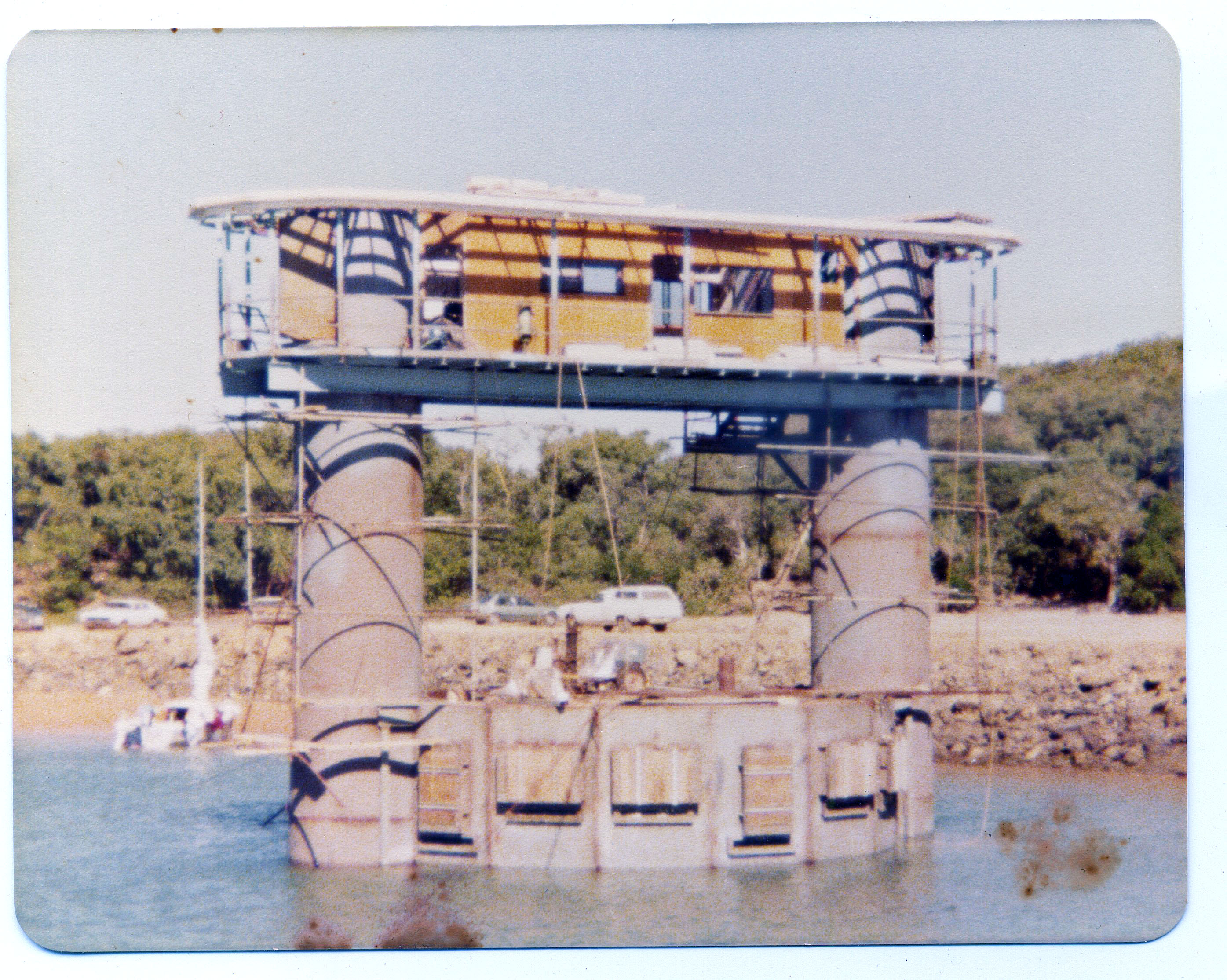 Estrutura de aço com dois grandes cilindros e molduras/escadas ao redor, sentado na água com parede de pedra e carros ao fundo 