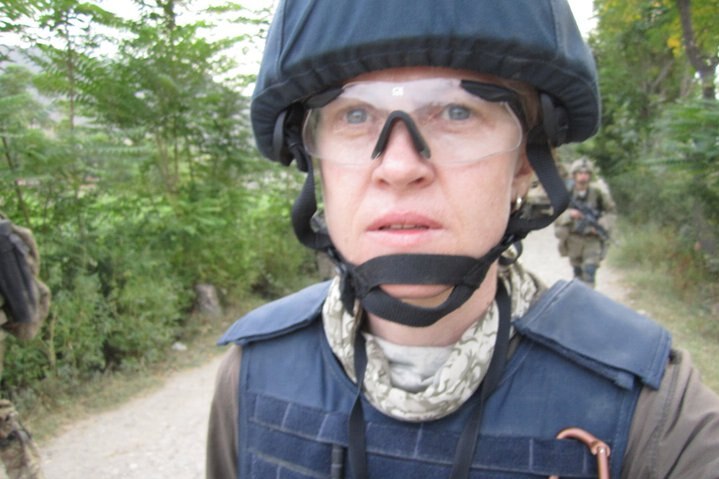 Sara wearing flak jacket, helmet and goggles walking down road with soldiers in background.