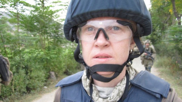 Sara wearing flak jacket, helmet and goggles walking down road with soldiers in background.