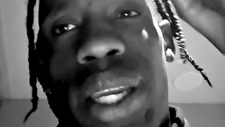 A close-up screenshot of rapper Travis Scott talking into a phone camera. The video has a black and white filter on it