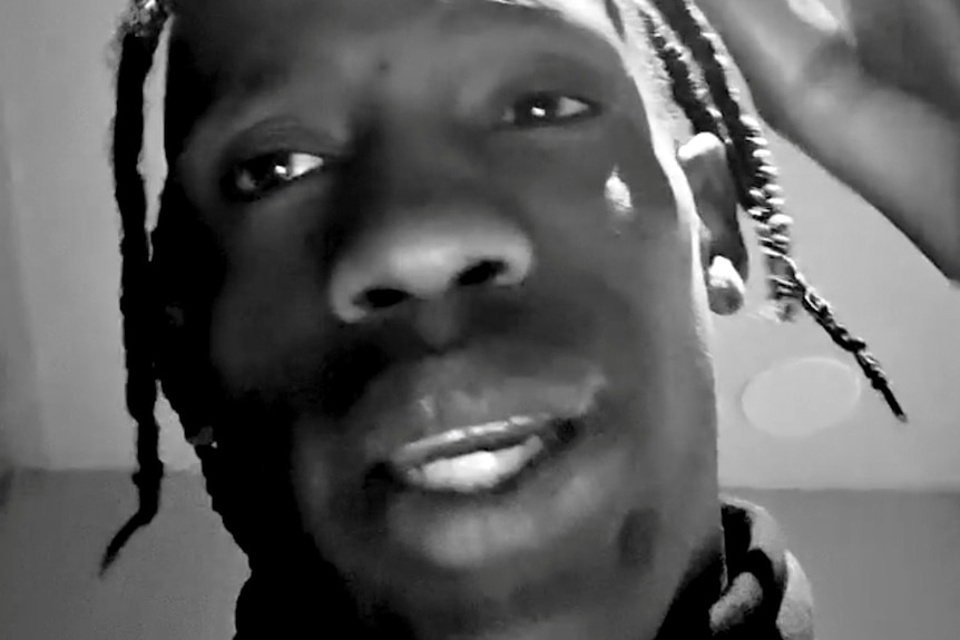 A close-up screenshot of rapper Travis Scott talking into a phone camera. The video has a black and white filter on it