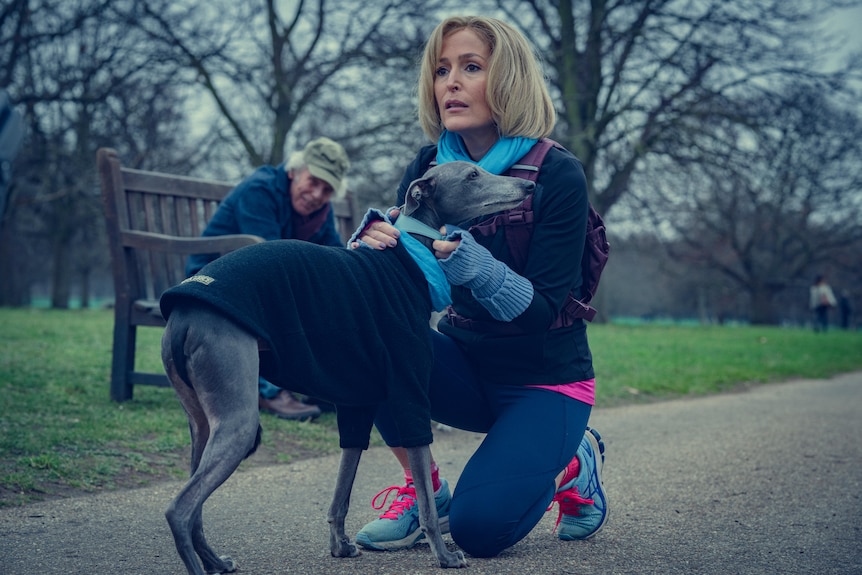 A film still of Gillian Armstrong in exercise gear, crouching down to hold a whippet dog. She is in a public park.