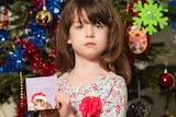 An unsmiling little girl holds up a Christmas card in front of a Christmas tree.