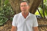 A man in his 50's in a white polo shirt, standing against trees