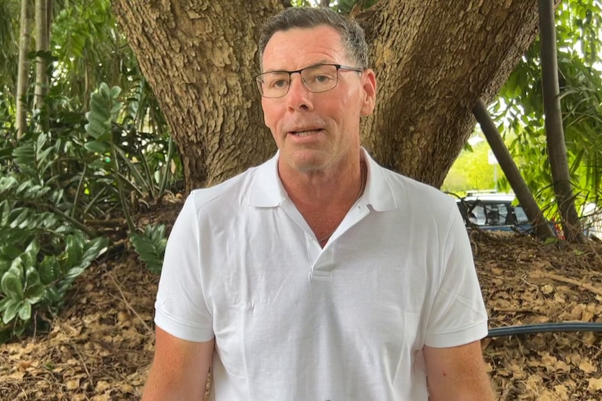 A man in his 50's in a white polo shirt, standing against trees