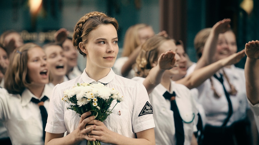 Colour still of Saskia Rosendahl wearing white military attire and holding bouquet of flowers in 2018 film Never Look Away.