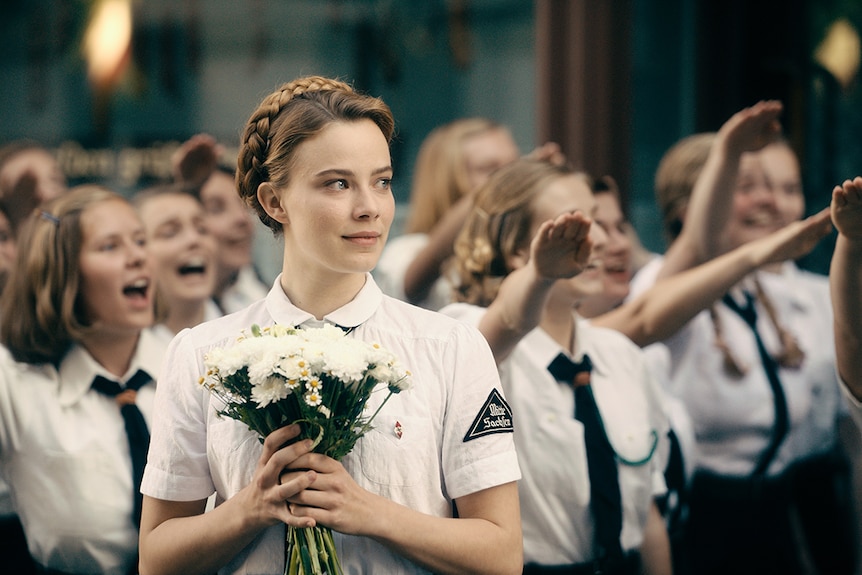 Colour still of Saskia Rosendahl wearing white military attire and holding bouquet of flowers in 2018 film Never Look Away.