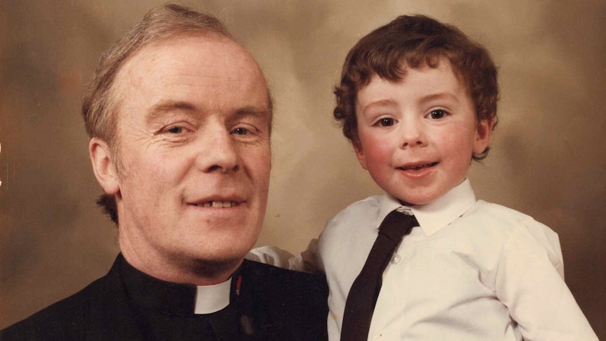 A young boy is held up by his father, who is wearing a priest's collar.