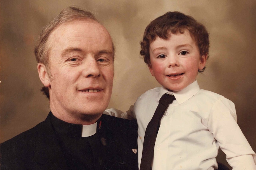 A young boy is held up by his father, who is wearing a priest's collar.