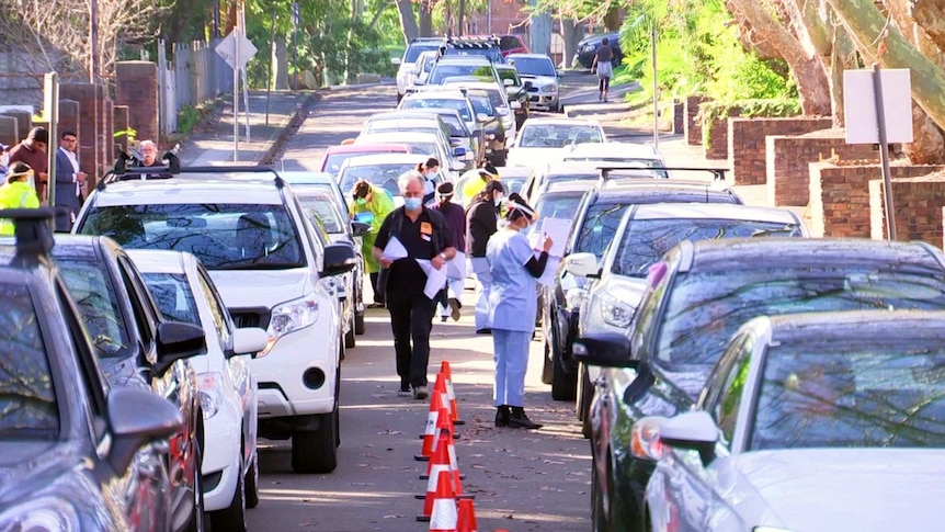 A row of cars lined up on a street with people standing next to them.