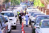 A row of cars lined up on a street with people standing next to them.