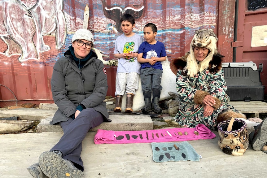 Daniel sitting on ground next to woman with wolf head hat and stone implements on rug and two children watching on.