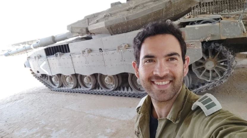 A young man in military uniform takes a selfie in front of a tank.