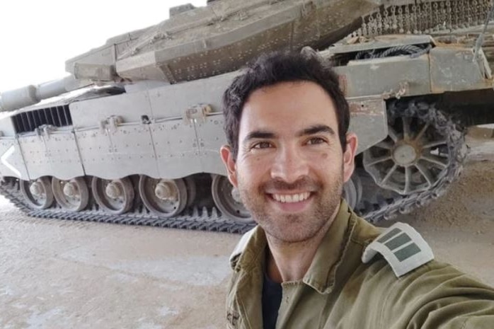 A young man in military uniform takes a selfie in front of a tank.