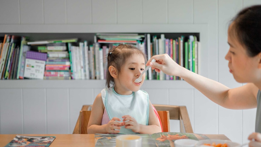 Mother feeding toddler in high chair with spoon sitting at table