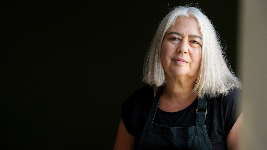 A woman wearing a t-shirt and apron sits as a window looking at the camera.