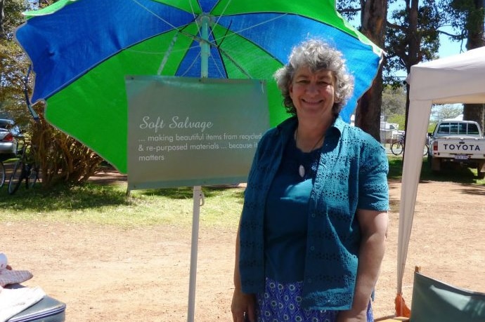 A woman in a green top stands next to a green and blue beach umbrella in a bush setting.