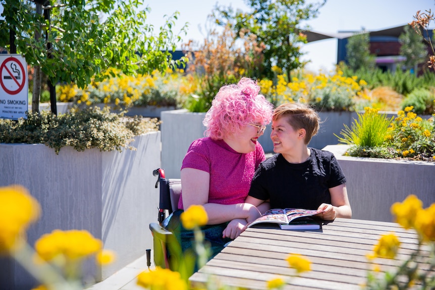 Ivy McGowan (left) sits next to Yoey Maxwell on a rooftop with plants behind them. They stare into each other's eyes and grin.