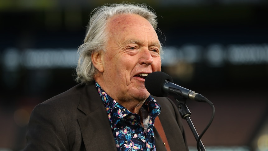 A close-up shot of a man in a floral shirt and jacket singing into a microphone.