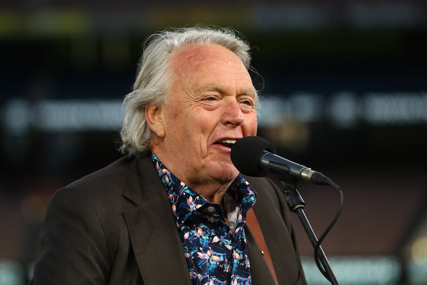 A close-up shot of a man in a floral shirt and jacket singing into a microphone.