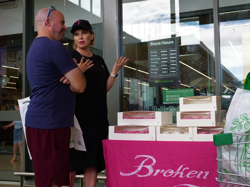 a man and woman stand near a cake stall, talking