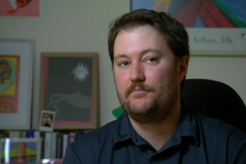 A man sits in a room in front of pictures on the wall