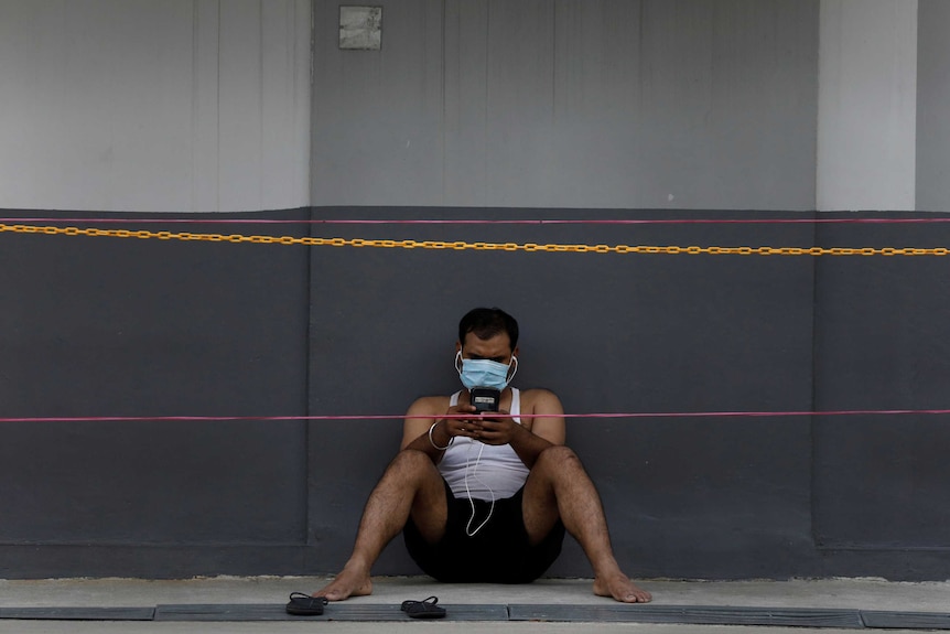 A man wears a face mask while sitting against a wall looking at his phone
