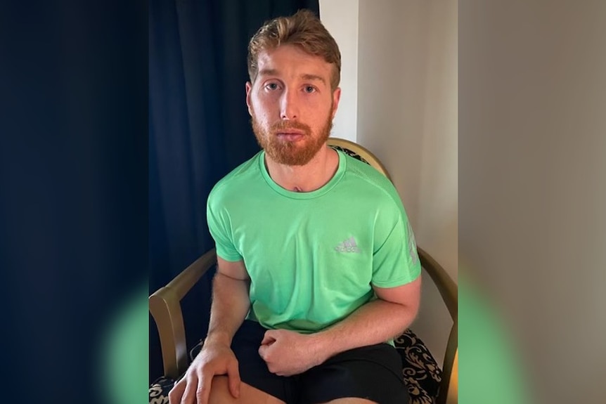 A blonde male in his twenties wears a green shirt and shorts as he sits in a chair