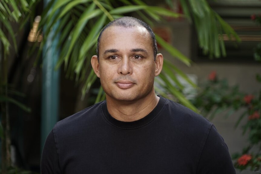 A man wearing a black shirt looks slightly past the camera with palm trees in the background. 
