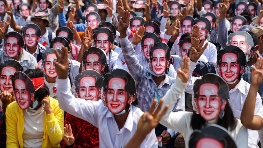 A large group of protesters wear masks depicting Aung San Suu Kyi, as they flash three-finger salutes.