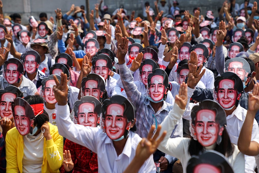 A large group of protesters wear masks depicting Aung San Suu Kyi, as they flash three-finger salutes.