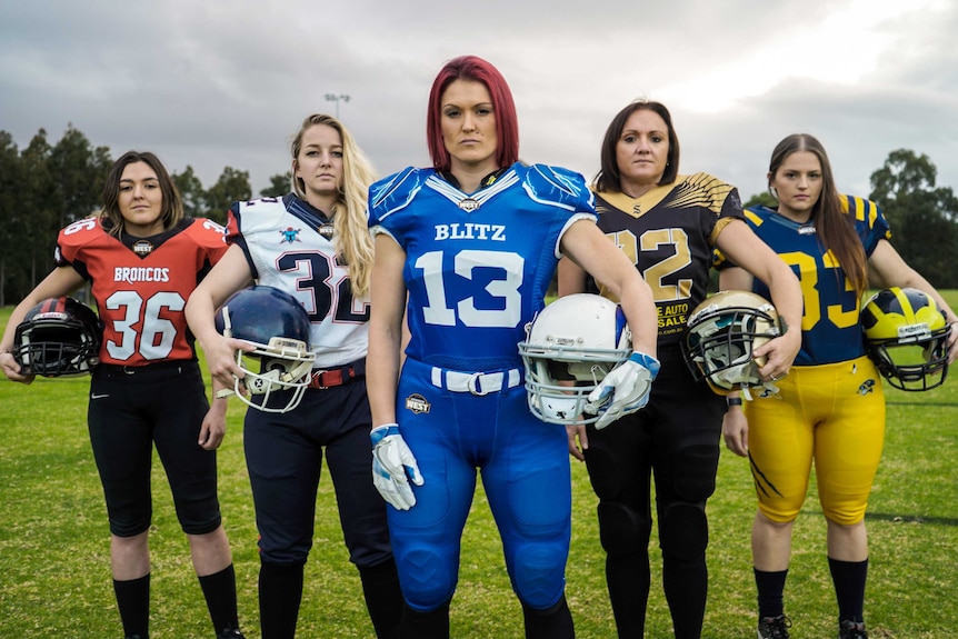Four women stand posing for a photo on grass wearing different gridiron uniforms and holding helmets.