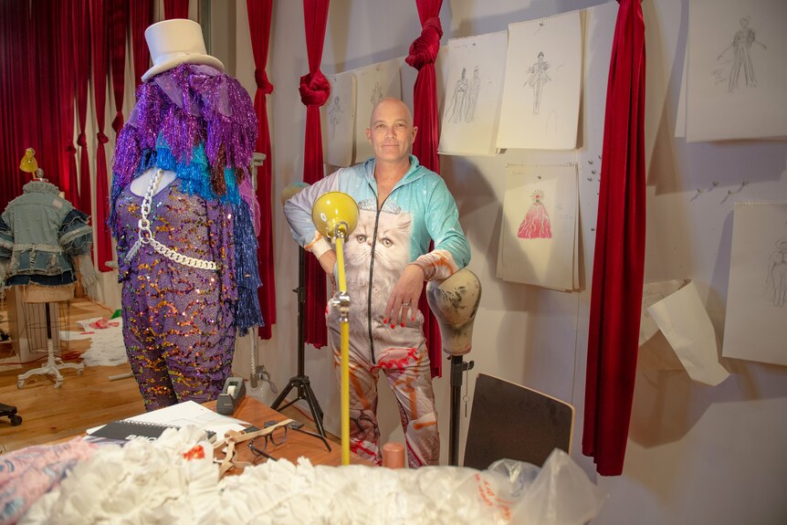 Taylor Mac, a white bald queer person dressed in a onesie with a cat printed on it, stands in a costume room.