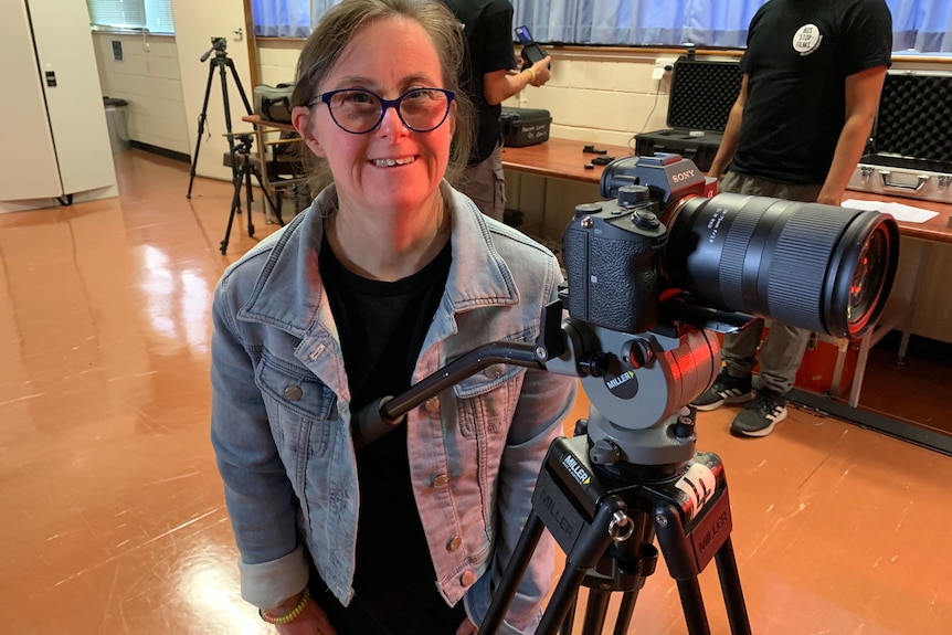 A dark haired woman smiles standing next to a camera on a tripod 