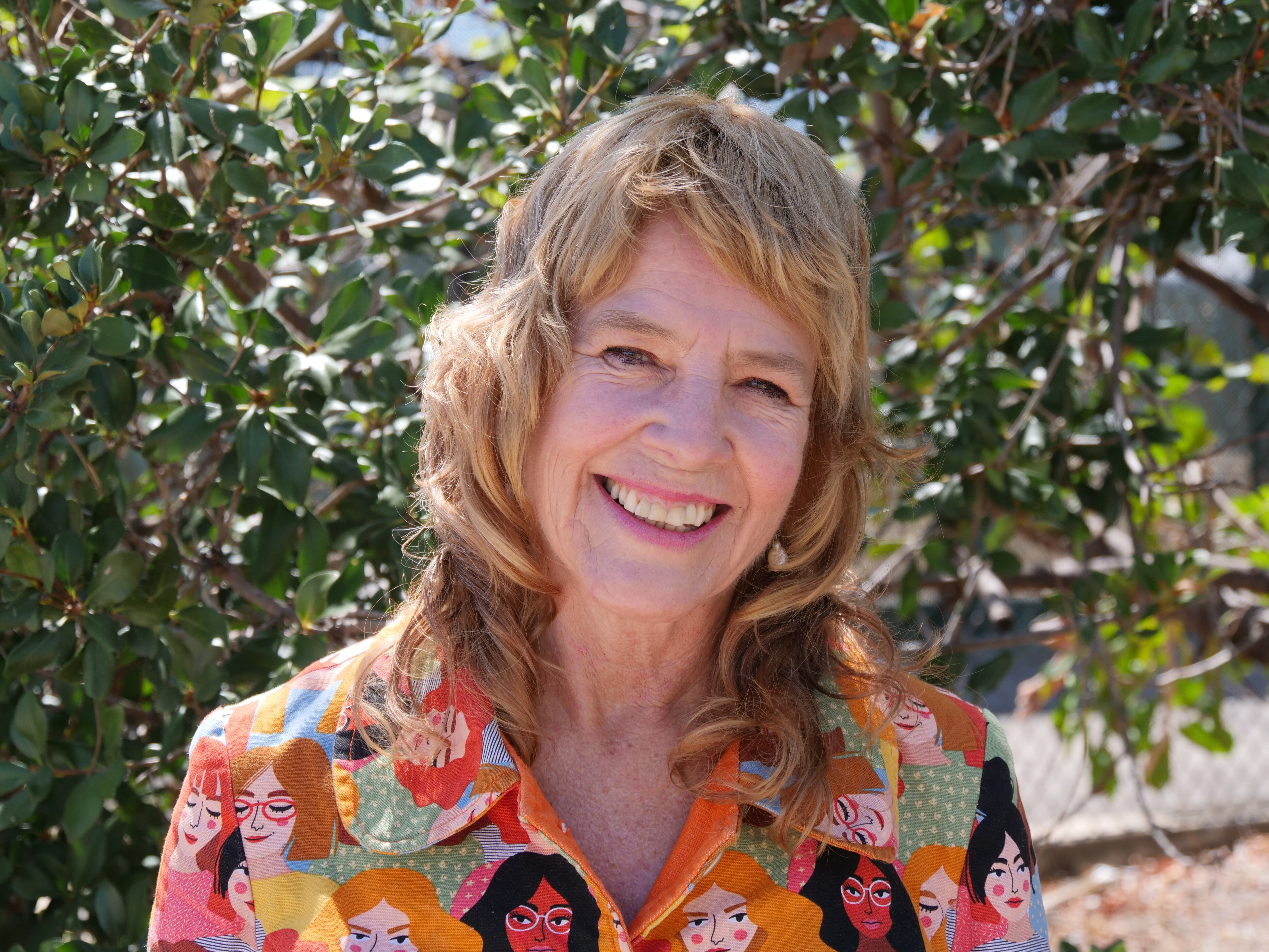 Uma mulher de meia-idade com cabelo encaracolado de comprimento médio sorri para a câmera em um vestido colorido. 