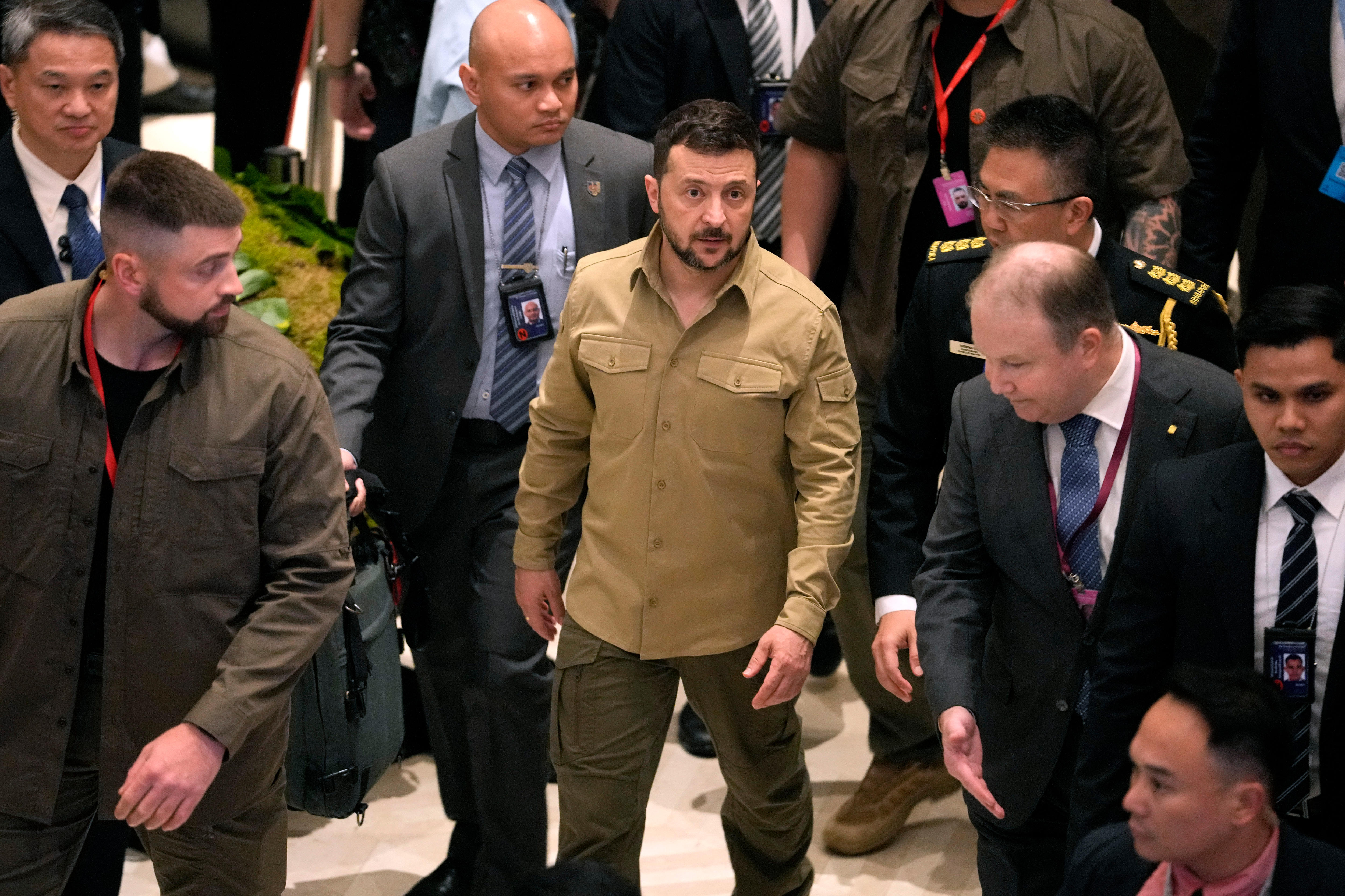 Volodymyr Zelenskyy leaves summit wearing a tan-coloured long sleeved shirt. He is surrounded by security guards