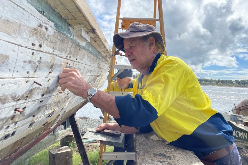 An older man wearing a work shirt and hat, works on the old timber hull of a 1940s workboat.