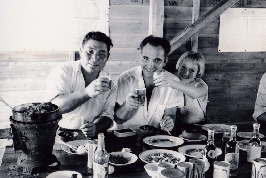 A Japanese worker raises his glass over lunch aboard a salvaged ship with a Territorian man and a young girl