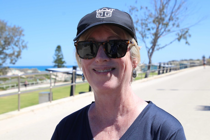 A head and shoulders shot of a woman wearing a cap and sunglasses near the beach.