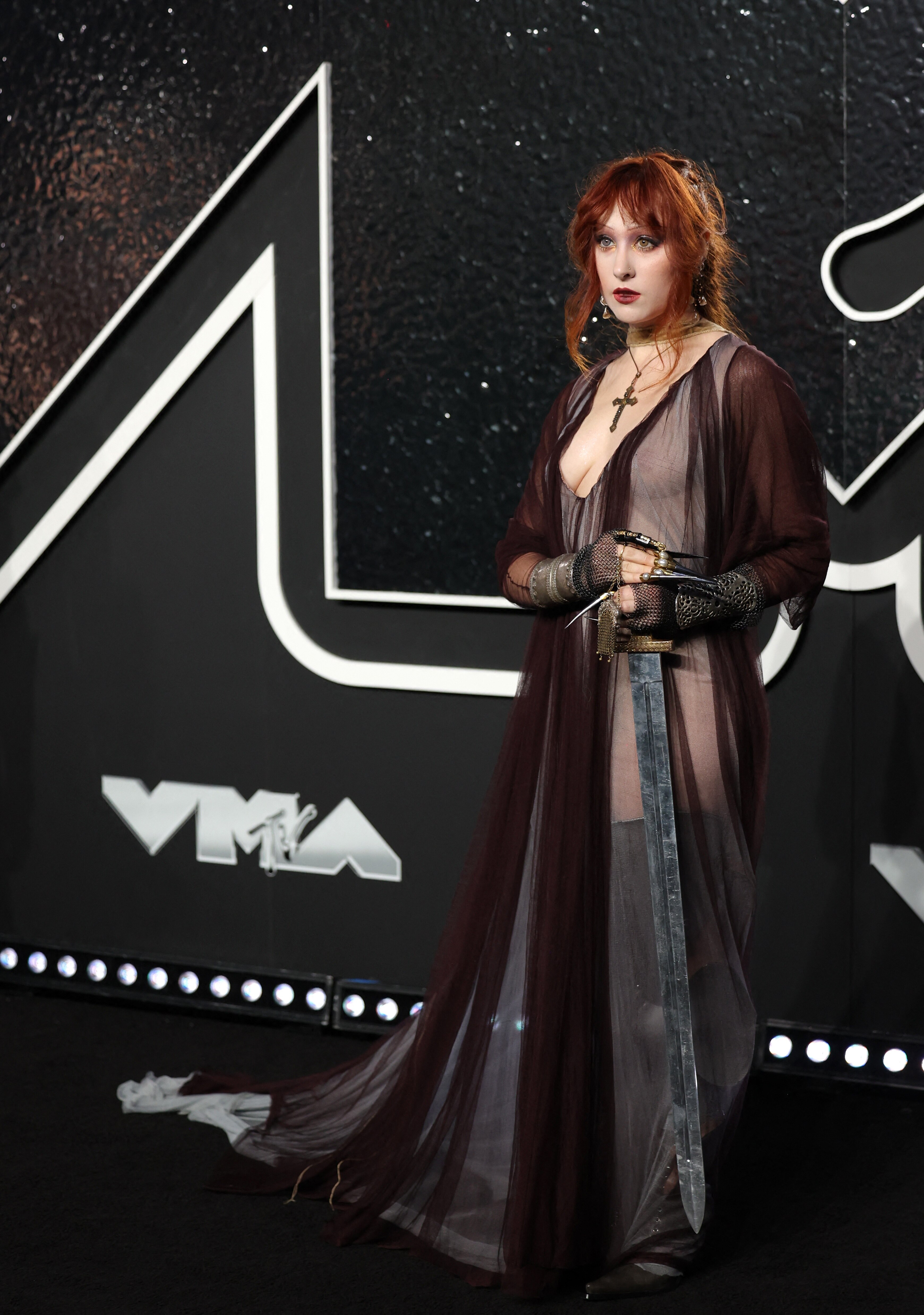 Chappell Roan on the VMAs red carpet wearing a black and maroon silky dress with cutouts. She also has very long fake nails